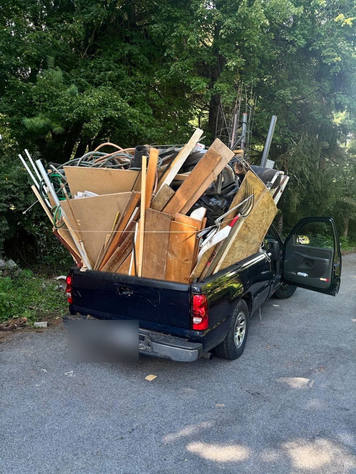 Black pickup at the curb with the door open, the bed piled with lumber, cardboard, hose, and yard debris.